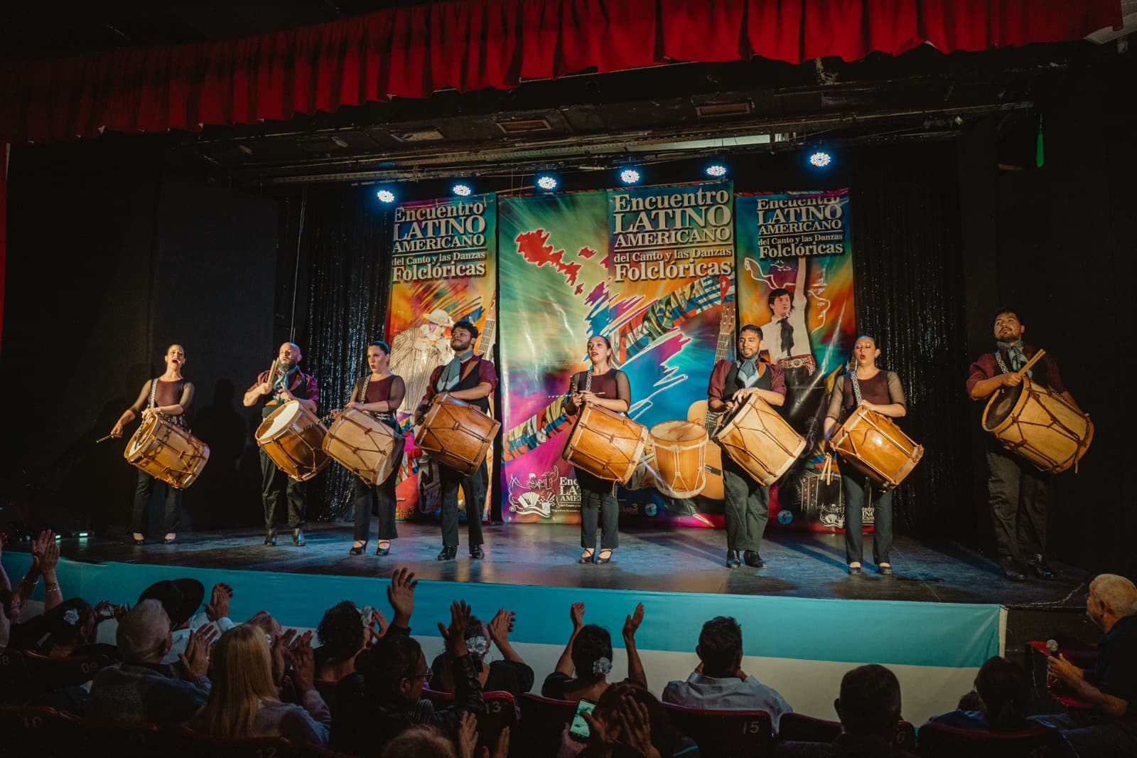Grupo de percusionistas tocando bombos legüeros en el escenario del Encuentro Latinoamericano con el público aplaudiendo